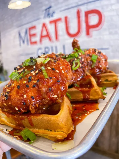 Spicy fried chicken served on waffles from The Meat Up Grill, topped with sesame seeds and green onions, offering a savory and sweet culinary delight.