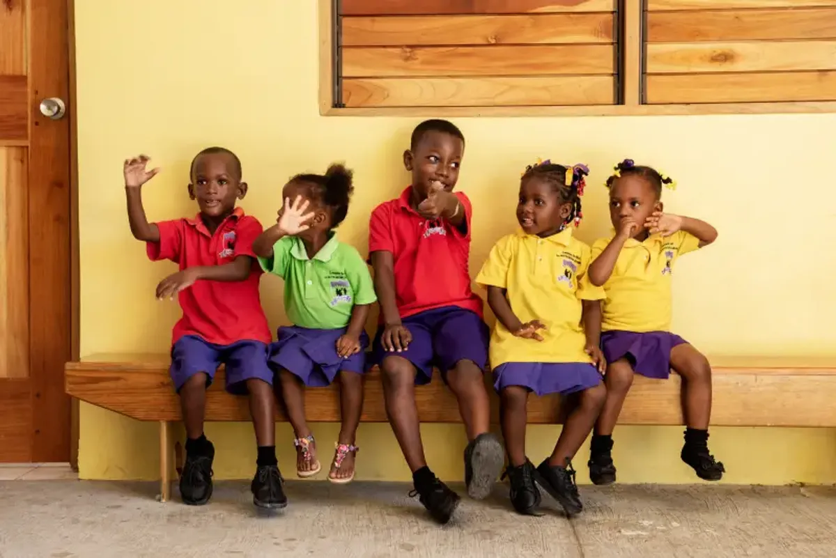 Group of young students from the Rockhouse Foundation school in Negril, Jamaica, sitting on a bench and waving at the camera—a sweet portrait of happy children.