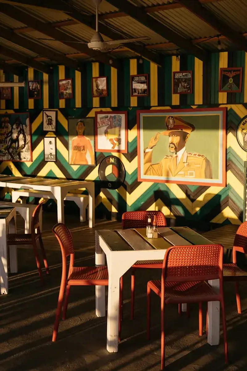 A casual, outdoor dining space with a vibrant, colorful striped wall in the red, green, and yellow of the Jamaican flag, featuring framed art and vintage posters, with white dining tables and orange chairs, capturing the laid-back, reggae-inspired vibe of the Rockhouse Hotel in Negril, Jamaica.
