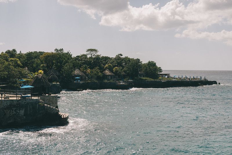 A panoramic view of the cliffside coastline at Rockhouse Hotel in Negril, Jamaica, with sparkling turquoise water, thatched-roof cabanas, and lush tropical greenery lining the shore.