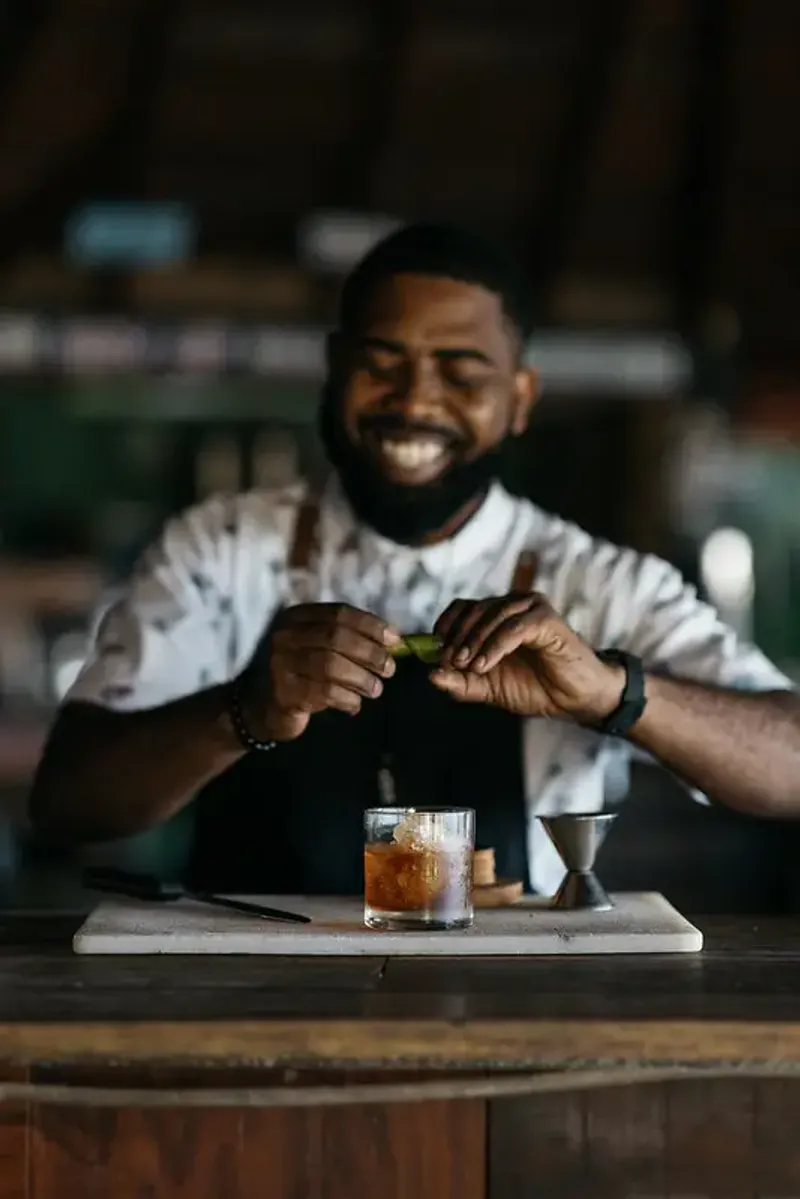 Smiling bartender garnishing a dark-colored cocktail with a lime twist at a bar in Rockhouse Hotel in Negril, Jamaica, showcasing the craft cocktail experience at Rockhouse Hotel.