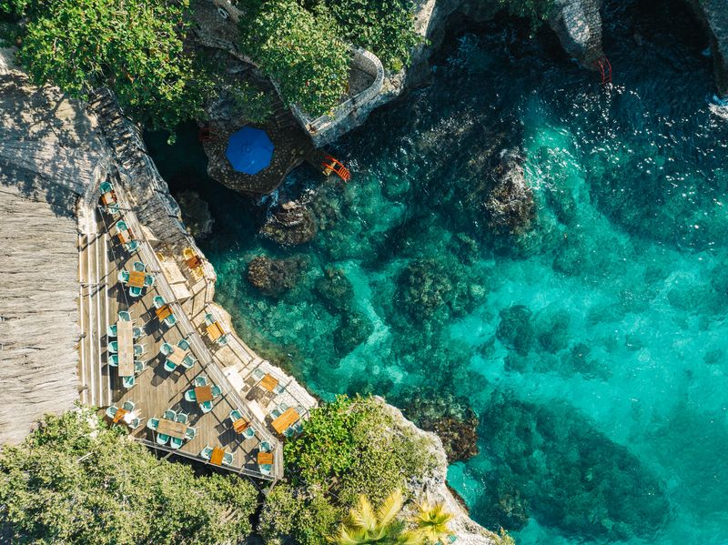 Aerial view of Rockhouse Hotel in Negril, Jamaica, showcasing a cliffside deck with turquoise seating overlooking crystal-clear Caribbean waters and rugged limestone formations.