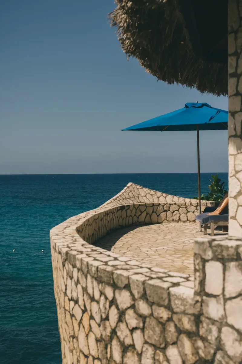 A scenic view of a stone balcony with a blue sun umbrella and lounge chair at Rockhouse Hotel in Negril, Jamaica, highlighting the resort's stunning cliffside views and private outdoor spaces.