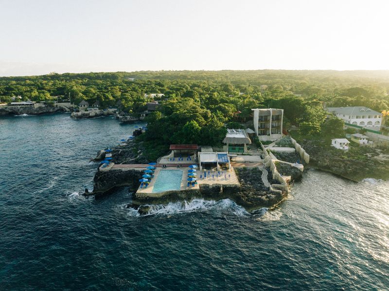 An aerial view of Rockhouse Hotel in Negril, Jamaica, perched on rugged cliffs above the Caribbean Sea, featuring a cliffside pool, sun loungers, and lush surrounding greenery.