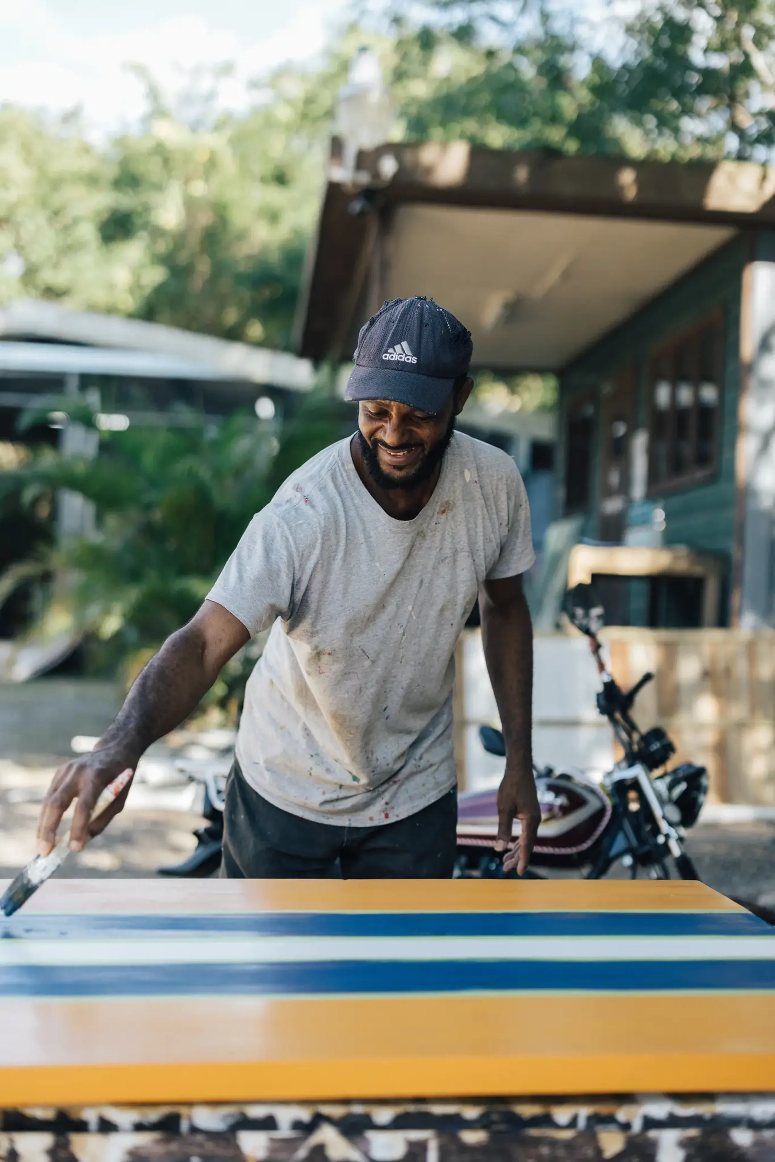 Man at Rockhouse Hotel in Negril, Jamaica, painting a table with vibrant stripes while a scooter is parked in the background—a glimpse of daily life.