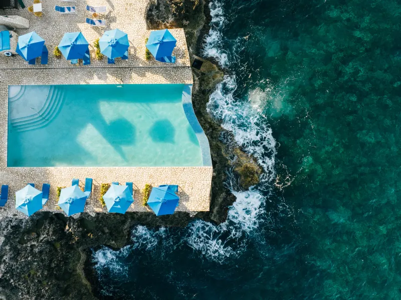 Aerial view of Catcha Falling Star's  vibrant turquoise infinity pool and lounge chairs on a cliffside terrace, with the Caribbean Sea in Negril, Jamaica, highlighting the resort's stunning oceanfront views and pool amenities.