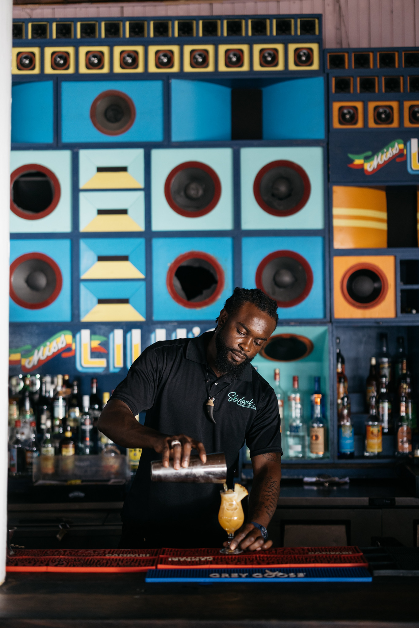 Bartender at Rockhouse Hotel in Negril, Jamaica, pouring a tropical yellow cocktail behind a vibrant, music-themed bar, framed by colorful oversized speaker boxes that create a lively, reggae-inspired backdrop.
