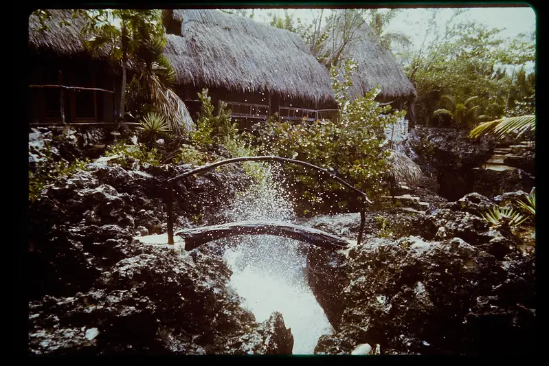 Rustic wooden bridge over a natural rock pool at Rockhouse Hotel in Negril, Jamaica, surrounded by lush tropical foliage and traditional thatched-roof villas, offering a serene and picturesque retreat