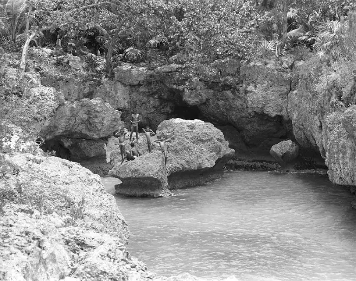 Vintage black-and-white photo of children playing on the rocky shoreline at Rockhouse Hotel in Negril, Jamaica, surrounded by lush tropical vegetation and natural rock formations