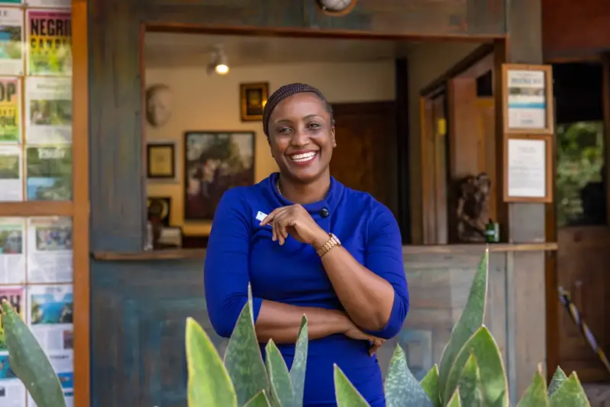 Smiling woman in a blue dress at the front desk of Rockhouse Hotel in Negril, Jamaica, standing in front of a rustic wooden counter—a portrait of a professional and friendly staff member.