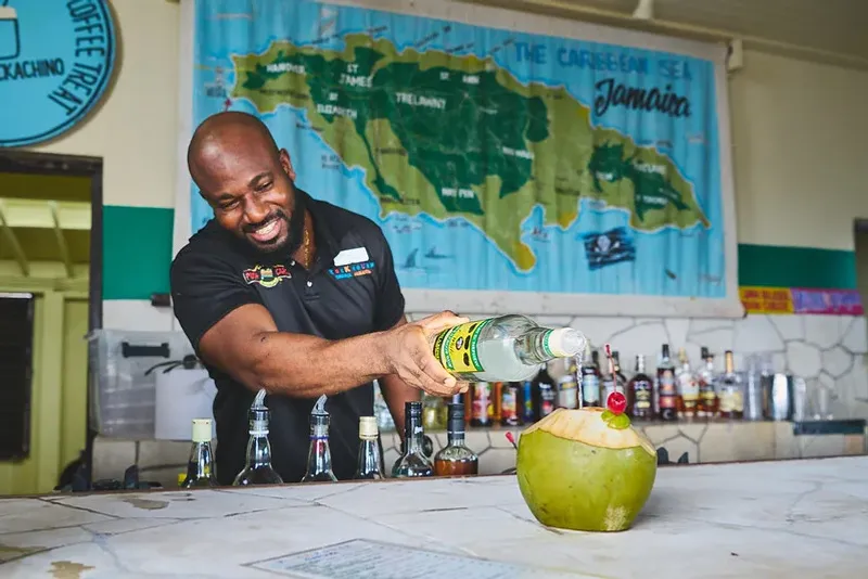 Bartender pouring a J Wray and Nephew white rum float into a coconut at the bar with a map of Jamaica behind him, at Rockhouse Hotel in Negril, Jamaica, showcasing the authentic bar experience at Rockhouse Hotel.