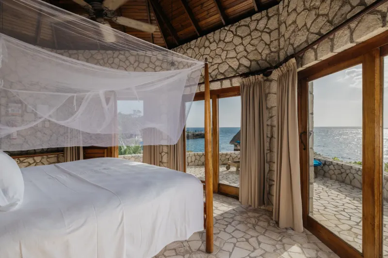 The interior of a thatched-roof villa at Rockhouse Hotel in Negril, Jamaica, featuring a comfortable bed with a white canopy and rustic wooden decor, highlighting the hotel's authentic and serene room design.