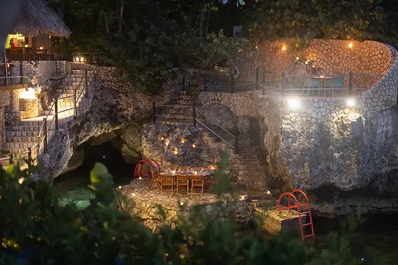 A cozy, candlelit outdoor table with two chairs, set on a rocky cove next to the water, showcasing the unique, natural architecture and scenic location of the Rockhouse Restaurant at Rockhouse Hotel in Negril, Jamaica.