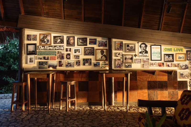 The warm and rugged indoor lounge at the Rockhouse Restaurant with natural stone walls and a wooden ceiling, featuring vintage reggae posters, a bookshelf with books and a record player, and a woven rocking chair with a vibrant, patterned cushion, capturing the casual, bohemian atmosphere of the Rockhouse Hotel in Negril, Jamaica.