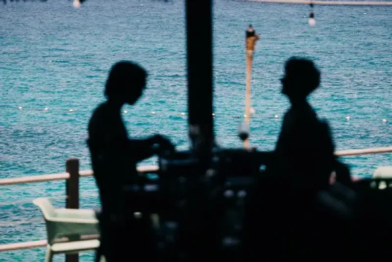 A blurred, silhouetted view of two people having a drink at the thatched-roof bar at the Rockhouse Restaurant, with the blue sea and a rope railing visible in the background, showcasing the unique, natural architecture and scenic location of Rockhouse Hotel in Negril, Jamaica.