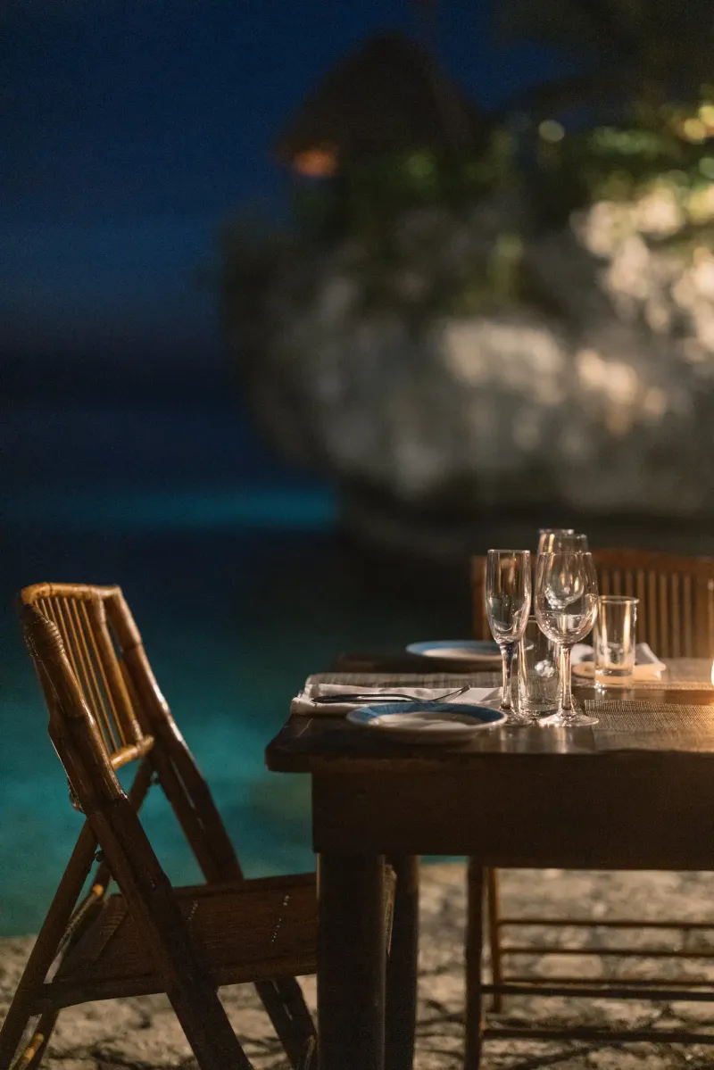 A cozy, rustic outdoor dining setting at Rockhouse Hotel in Negril, Jamaica, with a table set on a wooden deck overlooking the sea and lit by warm hanging lights, highlighting a romantic evening atmosphere.