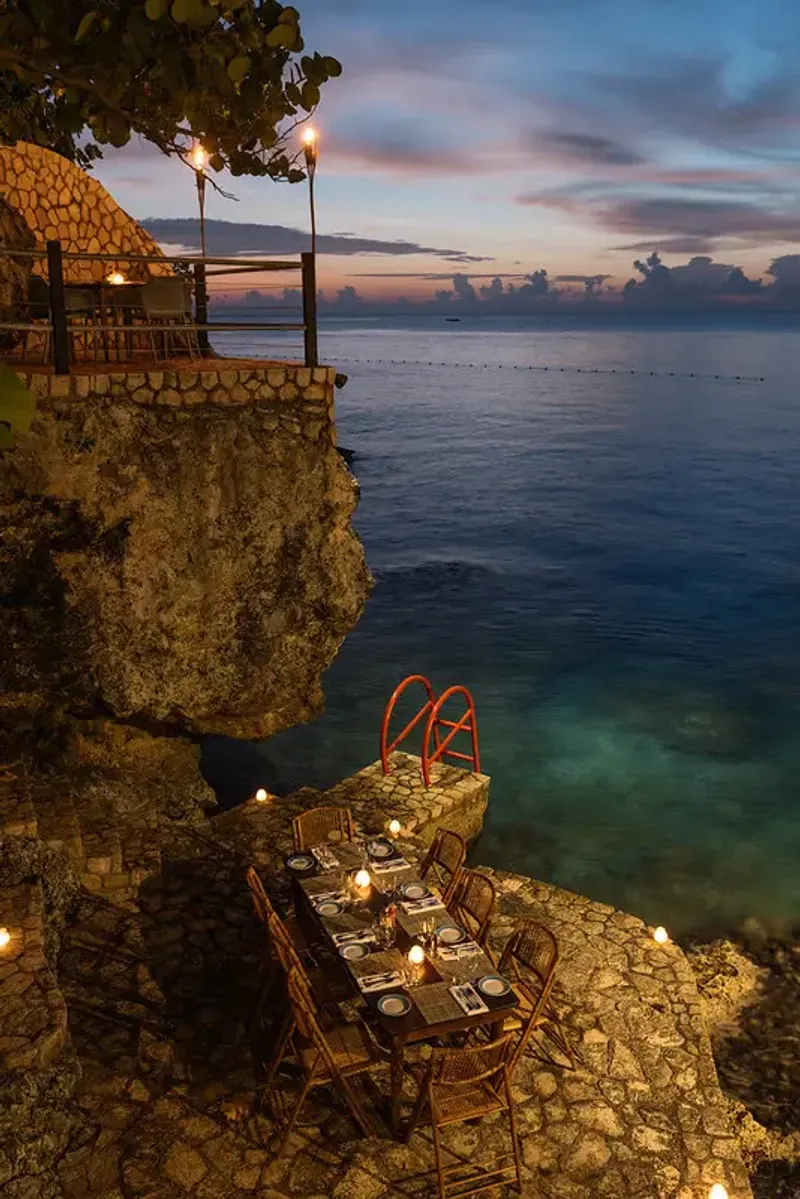 A romantic, candlelit dinner table with rattan chairs set on a rocky cliff overlooking the sea at sunset, showcasing the unique, natural architecture and scenic location of Rockhouse Restaurant at Rockhouse Hotel in Negril, Jamaica.