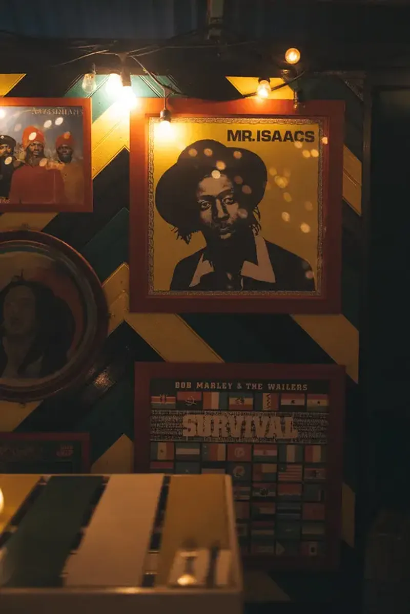 A close-up of a wall adorned with vintage reggae posters, including one of Bob Marley with a title that reads "MR. ISAACS" and another for "BOB MARLEY & THE WAILERS SURVIVAL," with a string of lights and a geometric striped pattern on the wall, highlighting the vibrant music theme of the Rockhouse Hotel in Negril, Jamaica.