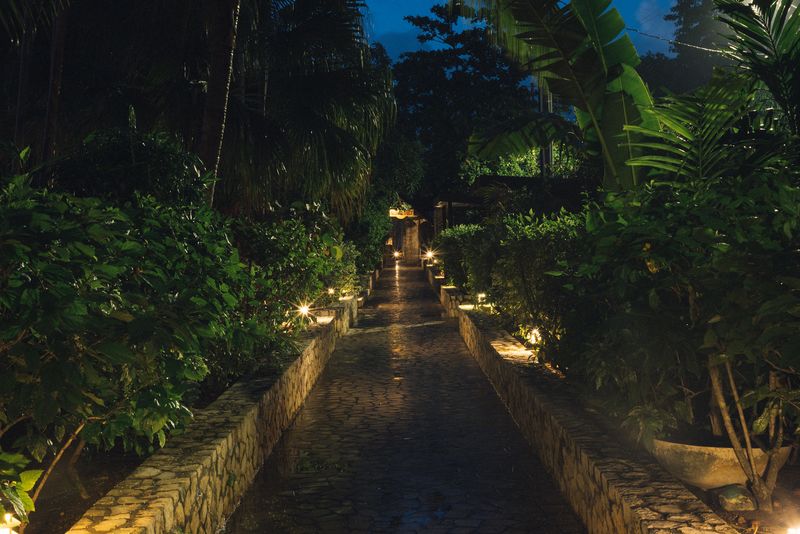 A softly lit stone pathway at Rockhouse Hotel in Negril, Jamaica, lined with dense tropical greenery and glowing lantern-style lights on a tranquil evening.
