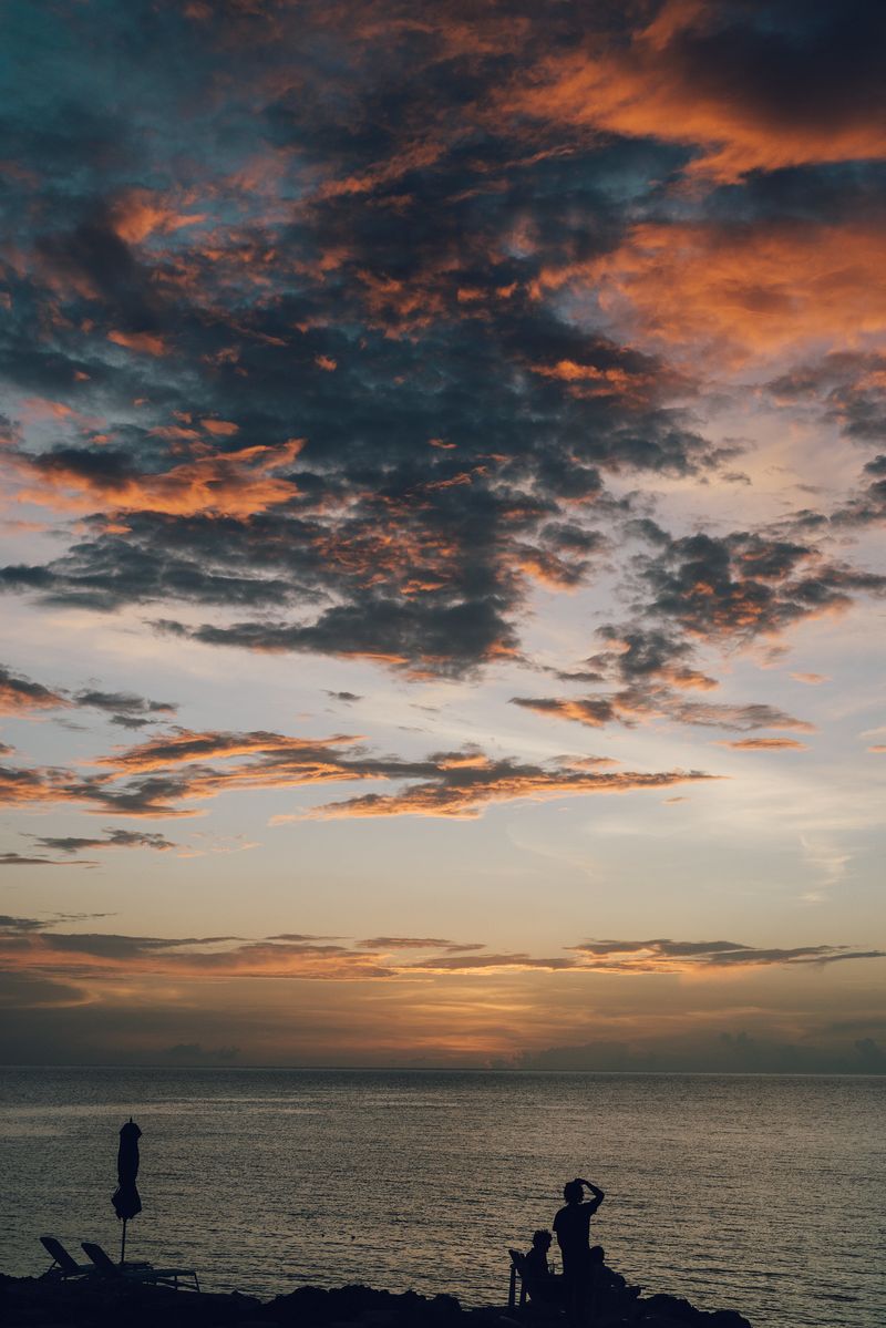 A dramatic sunset over the Caribbean Sea at Rockhouse Hotel in Negril, Jamaica, with silhouetted guests and lounge chairs along the cliffside as warm clouds glow overhead.