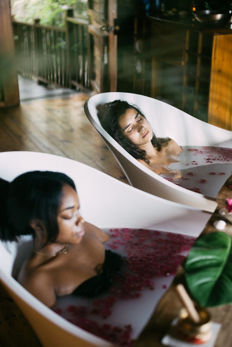 Two women enjoying a serene rose-petal bath ritual at Rockhouse Hotel in Negril, Jamaica, relaxing in modern soaking tubs surrounded by warm wood tones and tropical greenery.