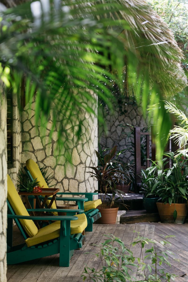 A peaceful veranda at Rockhouse Hotel in Negril, Jamaica, featuring green-and-yellow lounge chairs set against stone walls and surrounded by lush tropical plants.