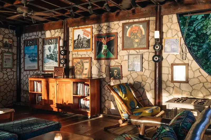 The warm and rugged indoor lounge at the Rockhouse Restaurant with natural stone walls and a wooden ceiling, featuring vintage reggae posters, a bookshelf with books and a record player, and a woven rocking chair with a vibrant, patterned cushion, capturing the casual, bohemian atmosphere of the Rockhouse Hotel in Negril, Jamaica.
