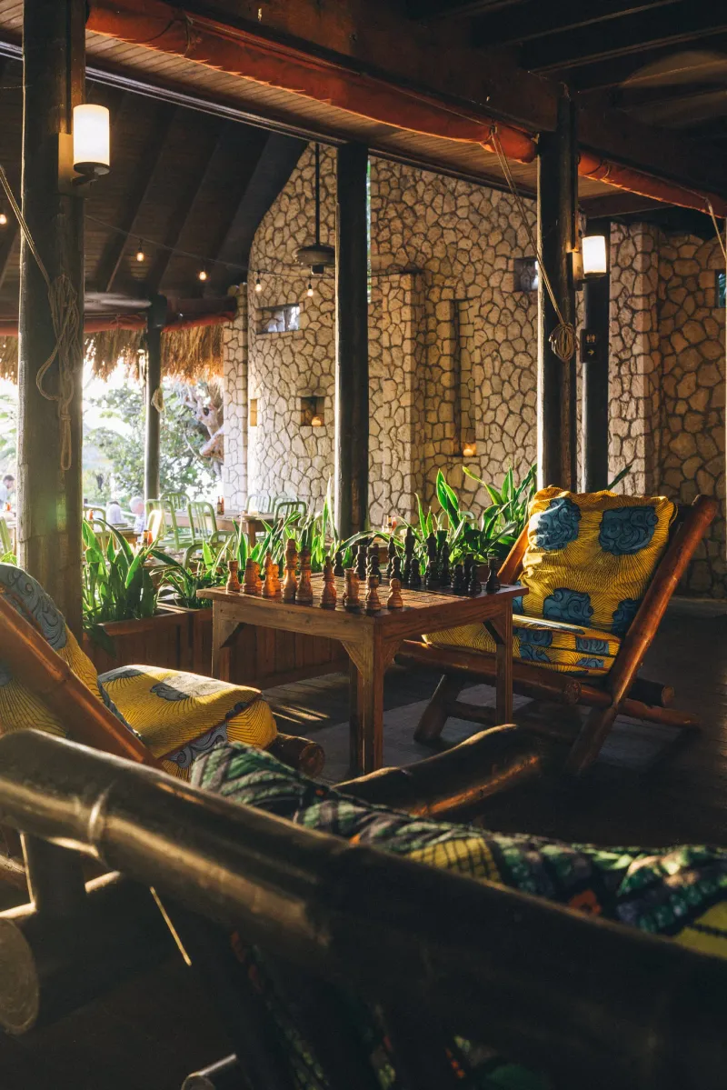 The warm and rugged indoor lounge at the Rockhouse Restaurant with natural stone walls and a wooden ceiling, featuring vintage reggae posters, a bookshelf with books and a record player, and a woven rocking chair with a vibrant, patterned cushion, capturing the casual, bohemian atmosphere of the Rockhouse Hotel in Negril, Jamaica.