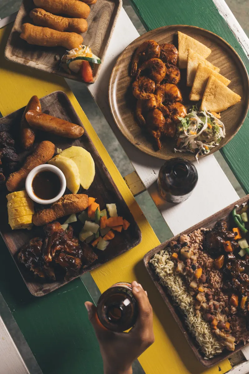 A platter of Jamaican food including rice and peas, curried chicken, and stewed beef, set on a counter in front of a colorful wall decorated with framed reggae album covers, highlighting the vibrant cultural and culinary experience at the Rockhouse Hotel in Negril, Jamaica.