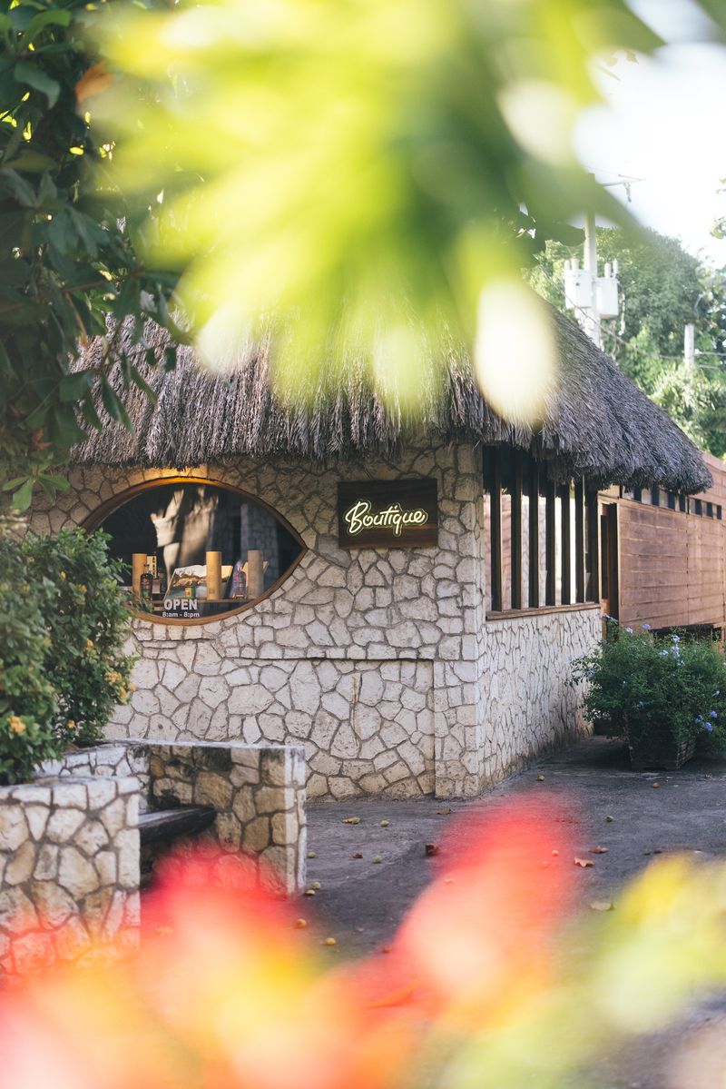 The Boutique at Rockhouse Hotel in Negril, Jamaica, housed in a rustic stone cottage with a thatched roof, framed by soft, colorful tropical foliage in the foreground.