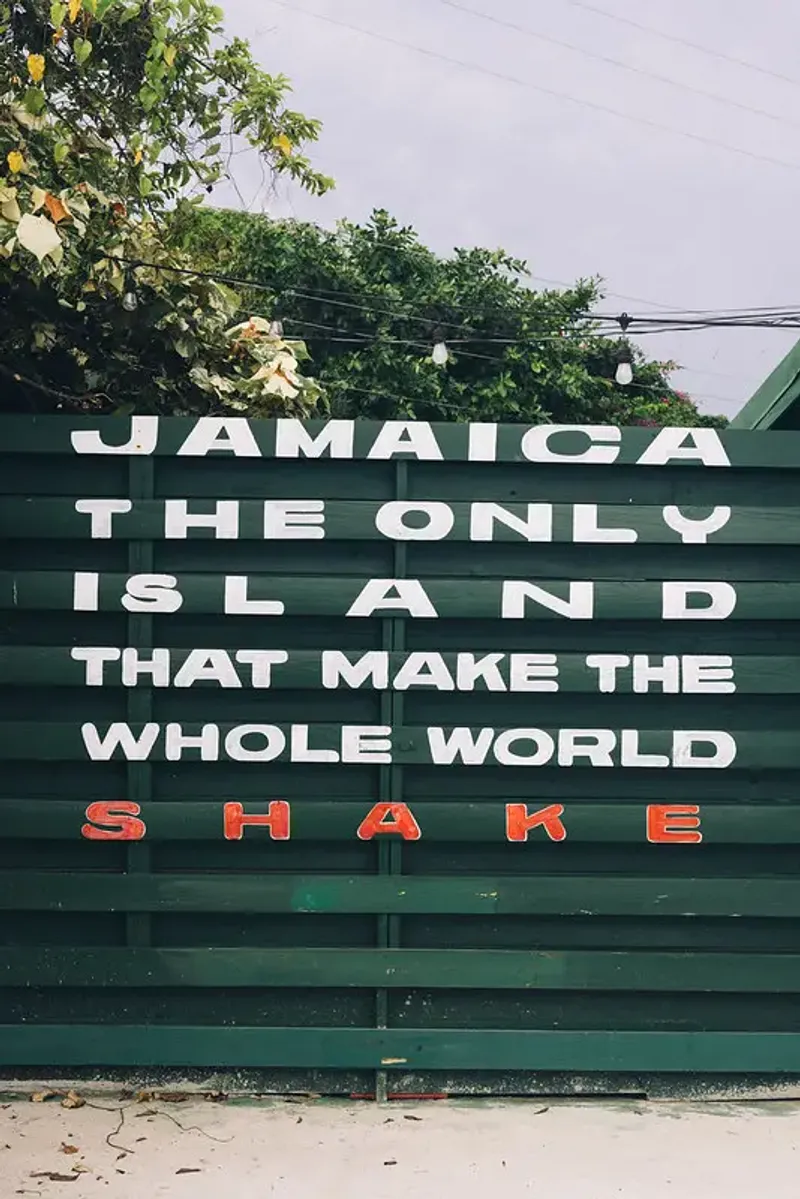 A wooden fence with a sign that reads "JAMAICA THE ONLY ISLAND THAT MAKE THE WHOLE WORLD SHAKE," at the Pushcart Rum Bar and Jerk Center featuring white and red lettering, highlighting the vibrant and influential cultural spirit of the Rockhouse Hotel in Negril, Jamaica.