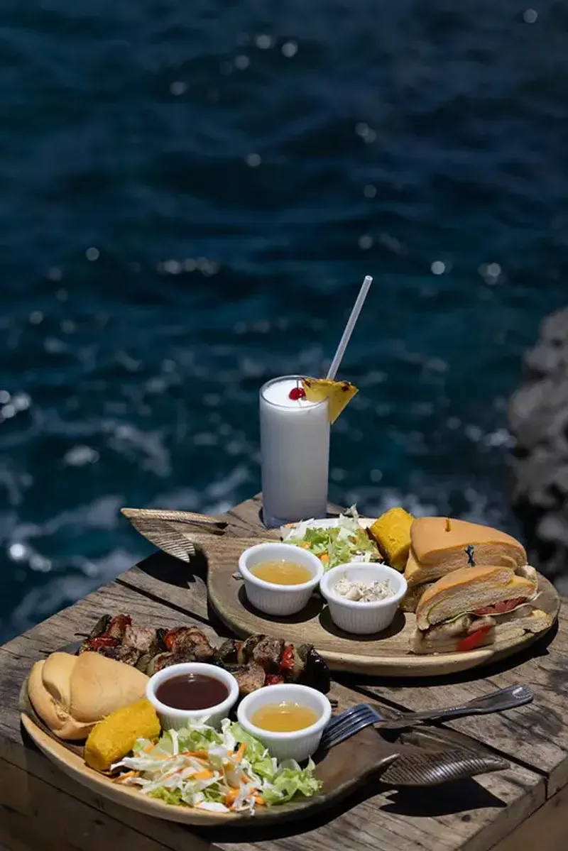 Platter of grilled skewers, sandwiches, and a tropical drink served at Rockhouse Restaurant in Negril, Jamaica, highlighting a flavorful lunch with a view at Rockhouse Hotel.