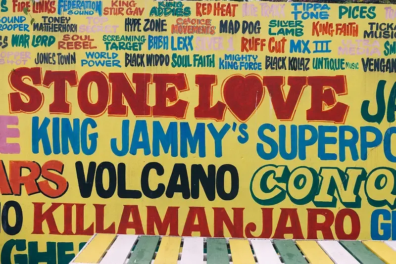 A colorful wall mural with bold, hand-painted reggae and dancehall names and phrases, including "STONE LOVE," "KING JAMMY'S SUPERPOWER," "VOLCANO," and "KILLAMANJARO," at the Pushcart Rum Bar and Jerk Center showcasing the deep connection to sound system culture at the Rockhouse Hotel in Negril, Jamaica.
