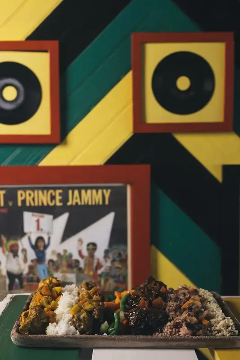 A platter of Jamaican food including rice and peas, curried chicken, and stewed beef, set on a counter in front of a colorful wall decorated with framed reggae album covers, highlighting the vibrant cultural and culinary experience at the Rockhouse Hotel in Negril, Jamaica.