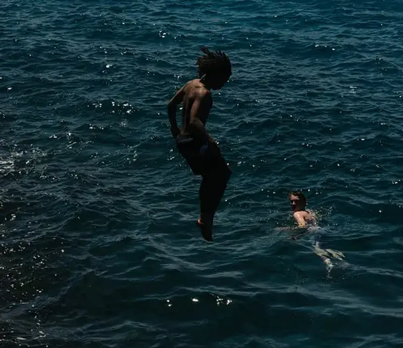 Man with dreadlocks leaping into the ocean with another person swimming nearby, at Rockhouse Hotel in Negril, Jamaica, showcasing cliff jumping into the natural coves at Rockhouse Hotel.