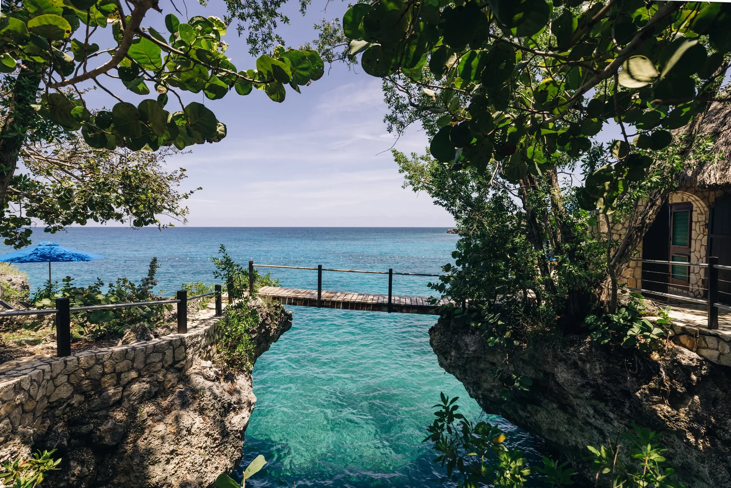 Scenic view at Rockhouse Hotel in Negril, Jamaica, featuring a turquoise cove framed by lush greenery and stone cliffs, with a rustic wooden bridge leading out toward the calm Caribbean Sea.