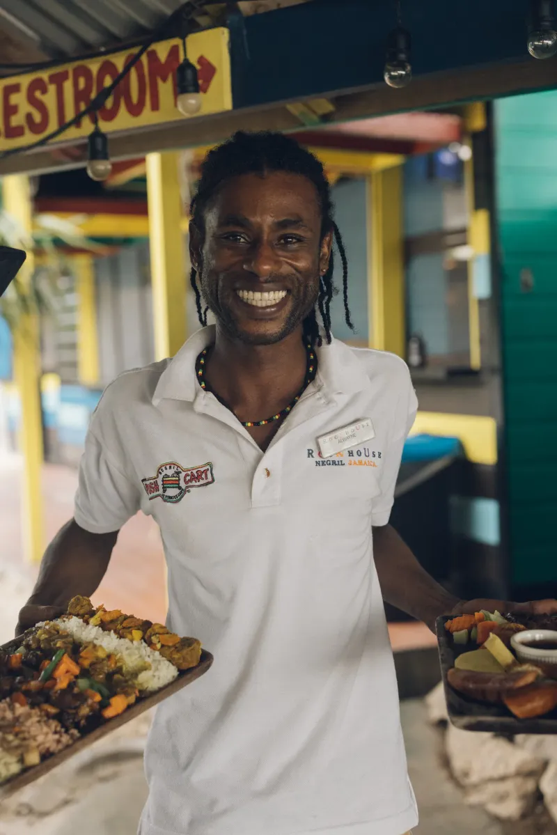 The smiling face of the award winning staff at Pushcart Jerk Center serving a platter of Jamaican food highlighting the vibrant cultural and culinary experience at the Rockhouse Hotel in Negril, Jamaica.