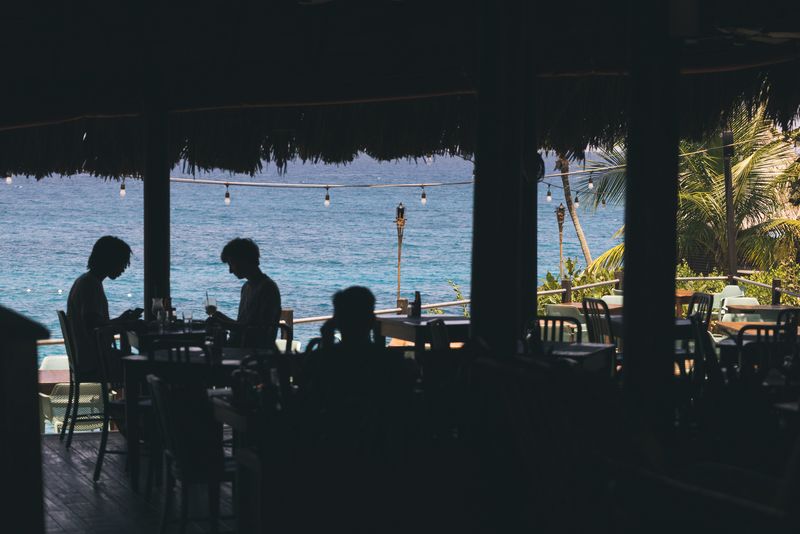 Guests dining in the shaded, open-air restaurant at Rockhouse Hotel in Negril, Jamaica, silhouetted against views of the turquoise Caribbean Sea.