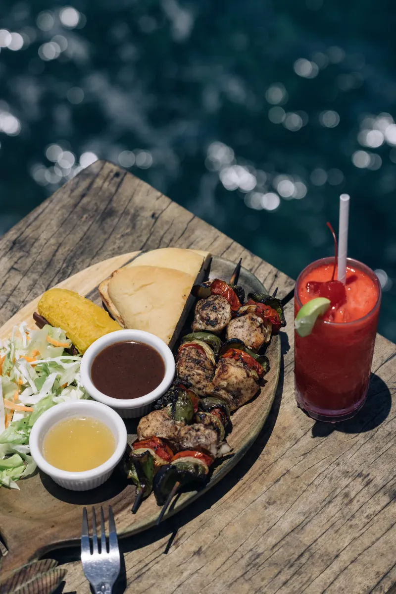 Platter of grilled skewers, sandwiches, and a tropical drink served at Rockhouse Restaurant in Negril, Jamaica, highlighting a flavorful lunch with a view at Rockhouse Hotel.