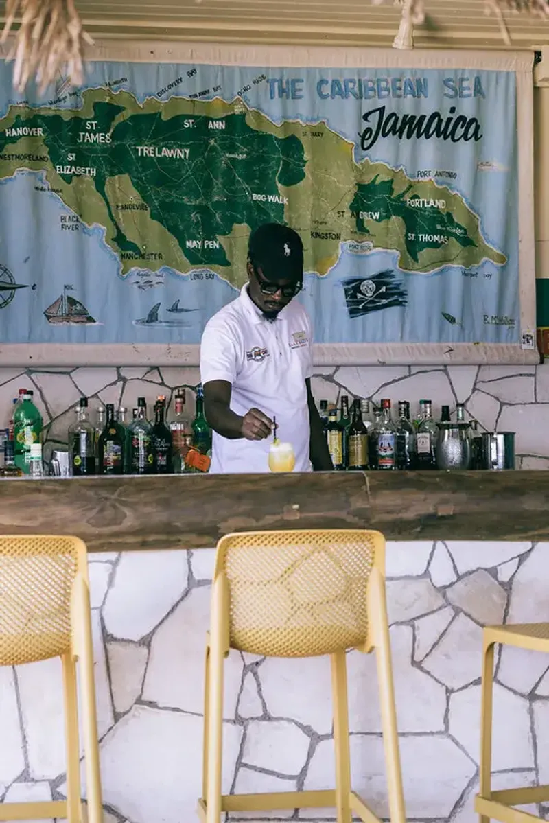 Bartender mixing a cocktail at the bar with a map of Jamaica behind him, at Rockhouse Hotel in Negril, Jamaica, showcasing the authentic bar experience at Rockhouse Hotel.