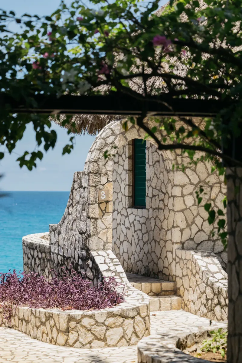 A vibrant view of a cliffside cabana with a lounge chair at Sunset at the Palms,  offering a picturesque spot to relax with the blue ocean and tropical foliage in Negril, Jamaica, highlighting the hotel's scenic relaxation areas.