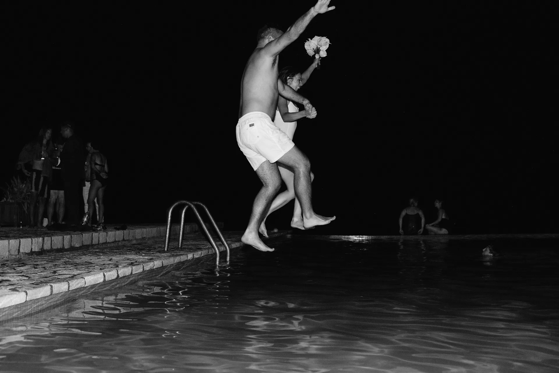 Guests at Rockhouse Hotel in Negril, Jamaica, jump hand-in-hand into the pool during a lively nighttime celebration, captured in a dynamic black-and-white moment.