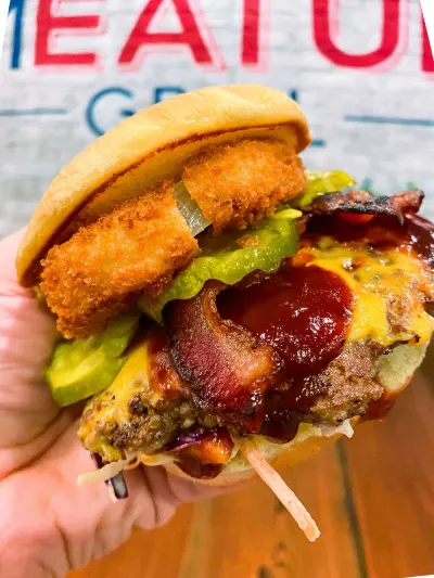 A mouthwatering burger from The Meat Up Grill, featuring crispy onion rings, bacon, pickles, and melted cheese, showcasing a delicious combination of flavors and textures.