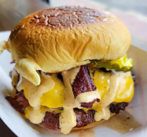 A juicy cheeseburger from The Meat Up Grill, topped with crispy bacon, pickles, and a generous drizzle of special sauce, highlighting a mouthwatering combination of flavors.