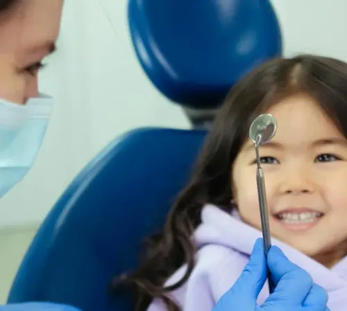 Smiling young girl sitting in a dental chair while a pediatric dentist shows her a dental mirror at East End Tooth Ferry in Southampton, New York.