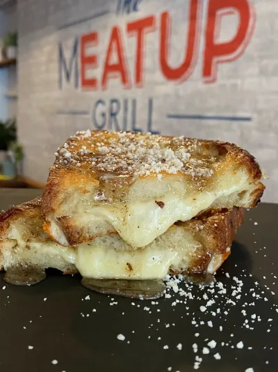 A gooey grilled cheese sandwich from The Meat Up Grill, with a crispy golden crust and melted cheese, set against the backdrop of the restaurant's logo.