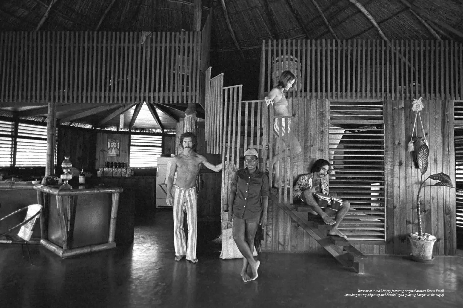 Historic interior of Awee-Maway, the original Rockhouse Hotel in Negril, Jamaica, featuring co-owner Erwin Finell and Frank Giglio playing bongos, capturing the resort's early bohemian spirit and unique thatched-roof design.