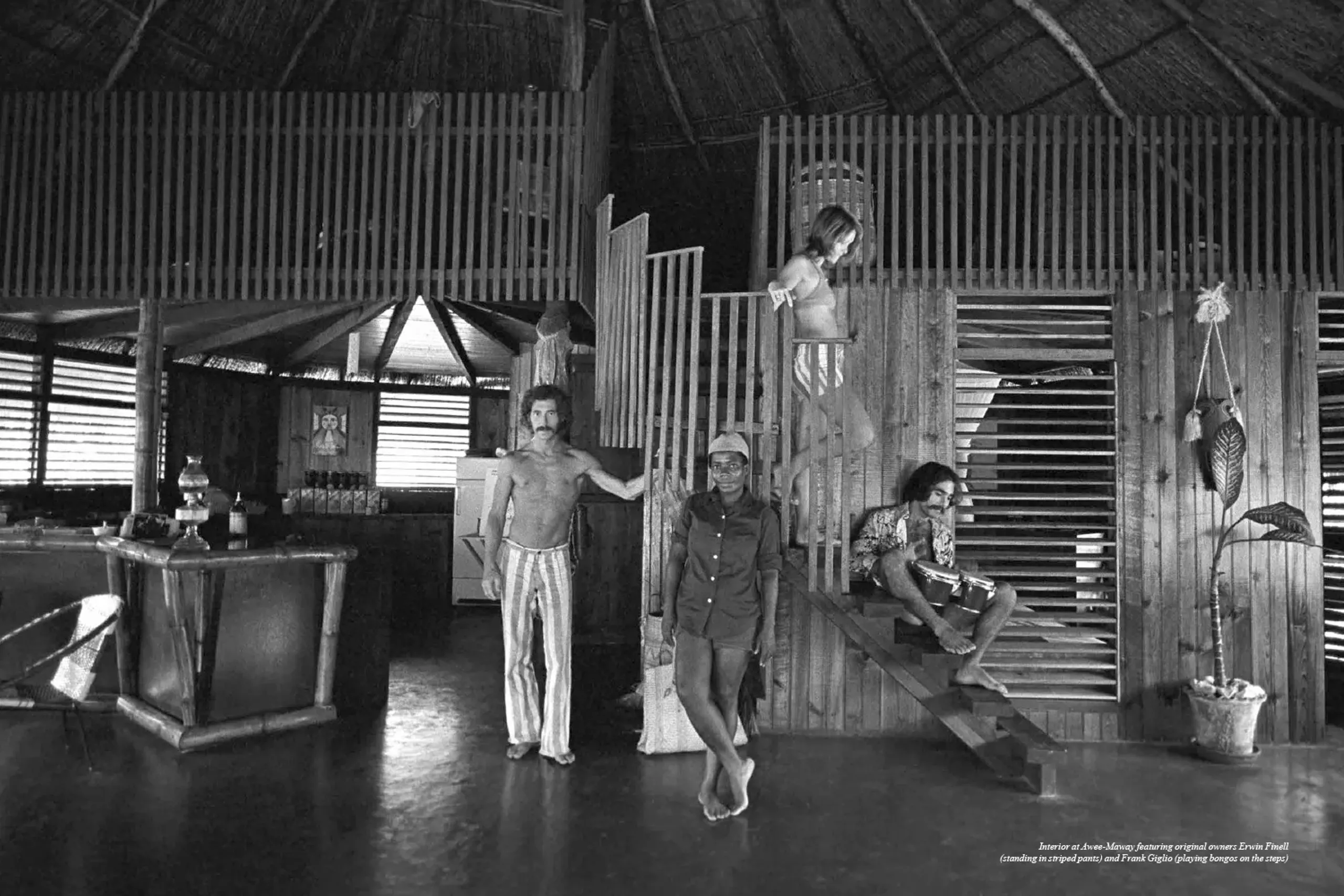 Historic interior of Awee-Maway, the original Rockhouse Hotel in Negril, Jamaica, featuring co-owner Erwin Finell and Frank Giglio playing bongos, capturing the resort's early bohemian spirit and unique thatched-roof design.