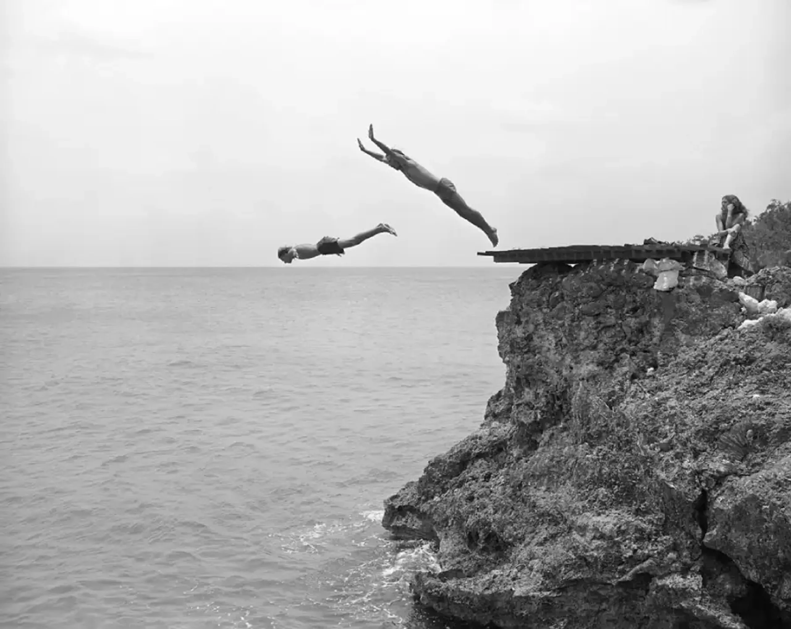 Vintage black-and-white photo of a guest diving off the cliffs at Rockhouse Hotel in Negril, Jamaica, with thatched-roof villas and lush tropical surroundings, capturing the resort's adventurous spirit and natural beauty