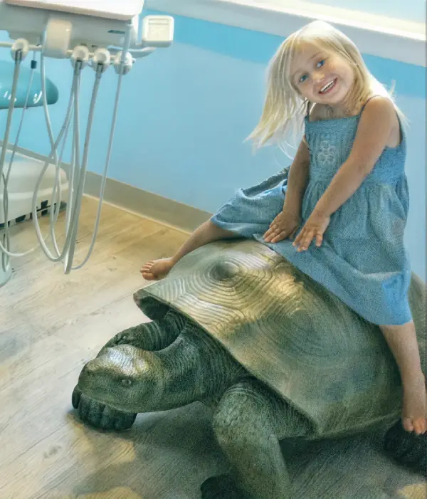 Smiling young girl sitting on a large turtle statue inside East End Tooth Ferry pediatric dental office in Southampton, New York